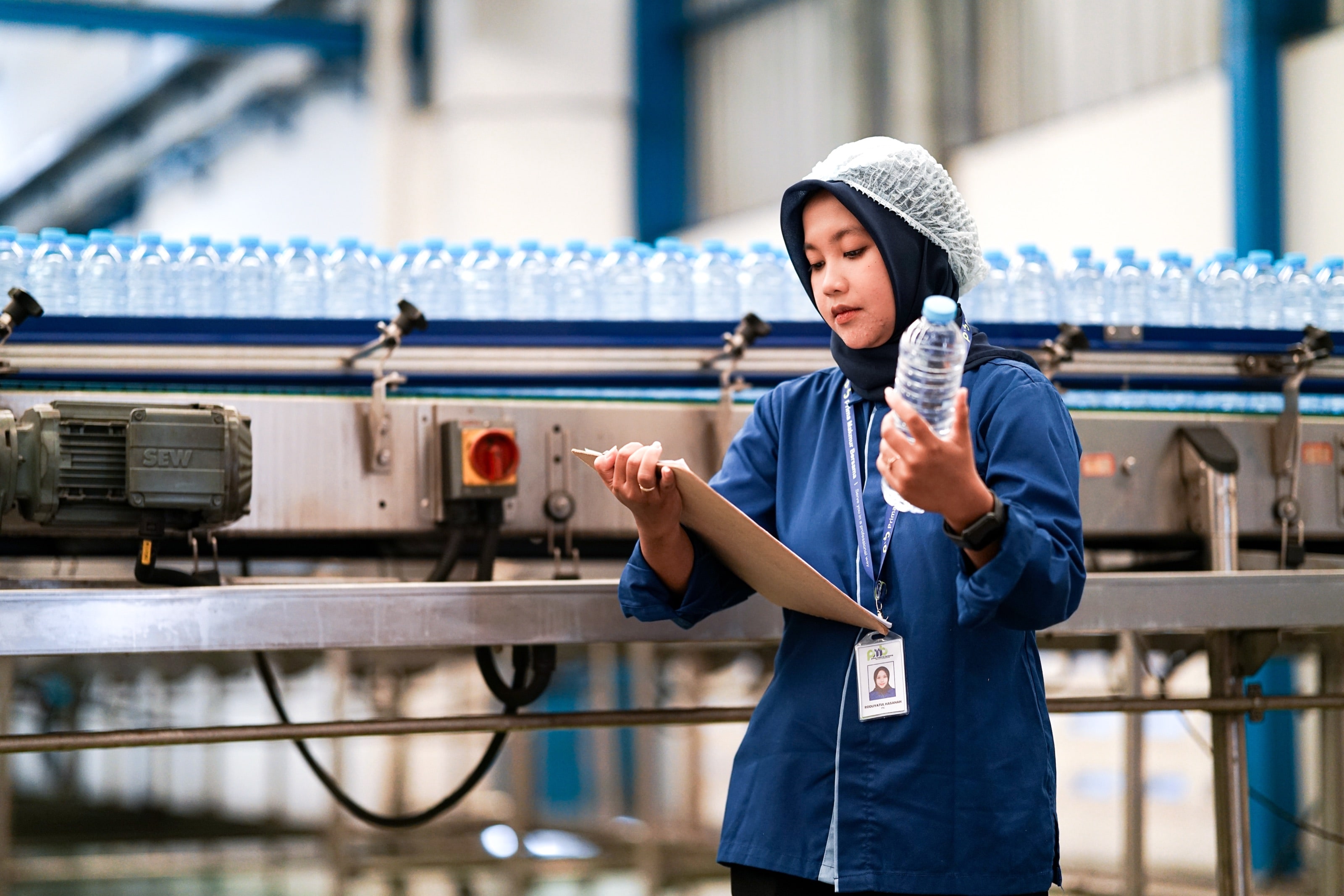 A PMB worker inspecting a bottle on a packaging line while recording on a clipboard
